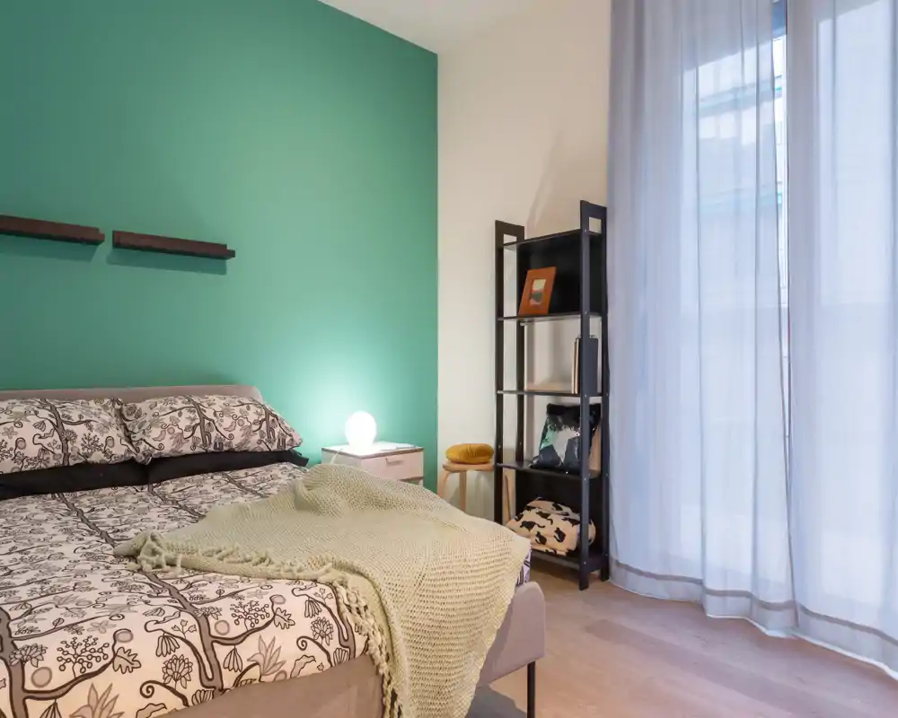 Dormitorio luminoso y decorado con cama doble estampada, pared verde de acento, lámpara de mesita y cortinas que filtran la luz natural.