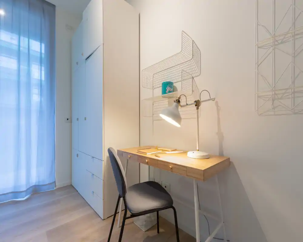 Rincón de trabajo luminoso y minimalista con escritorio de madera, silla y lámpara junto a un armario blanco alto y una cortina traslúcida — perfecto para un estudio o zona de trabajo en dormitorio.