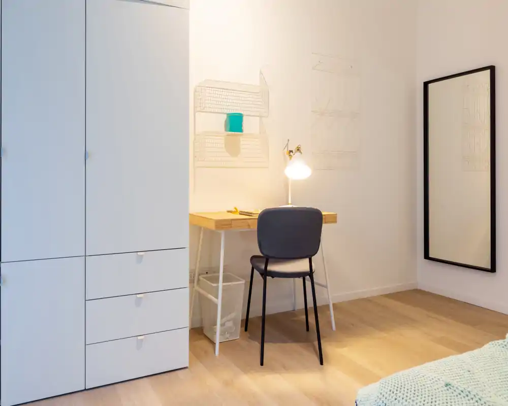 Rincón de dormitorio luminoso y minimalista con escritorio, silla, armario y lámpara; una pequeña parte de la cama se ve en el borde del encuadre.