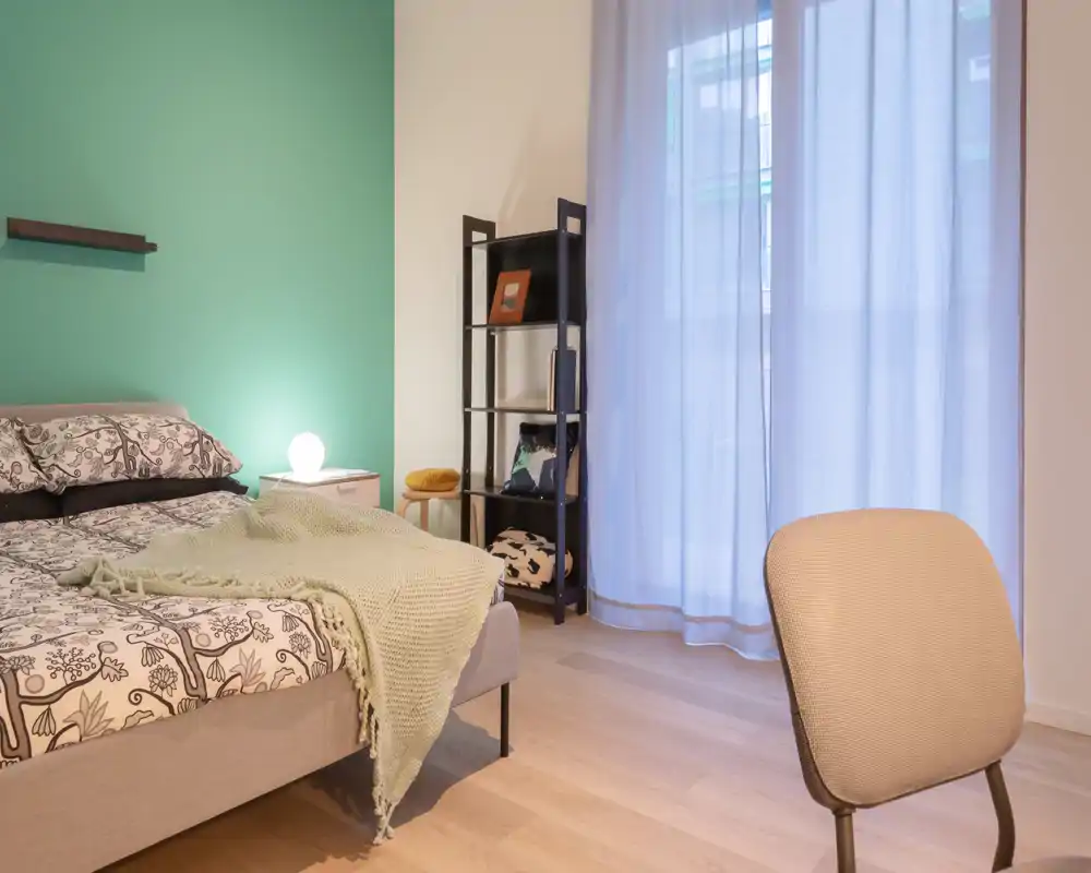 Dormitorio luminoso y bien decorado con cama estampada, manta suave y lámpara de mesita; gran ventana con cortinas que difunden la luz natural.