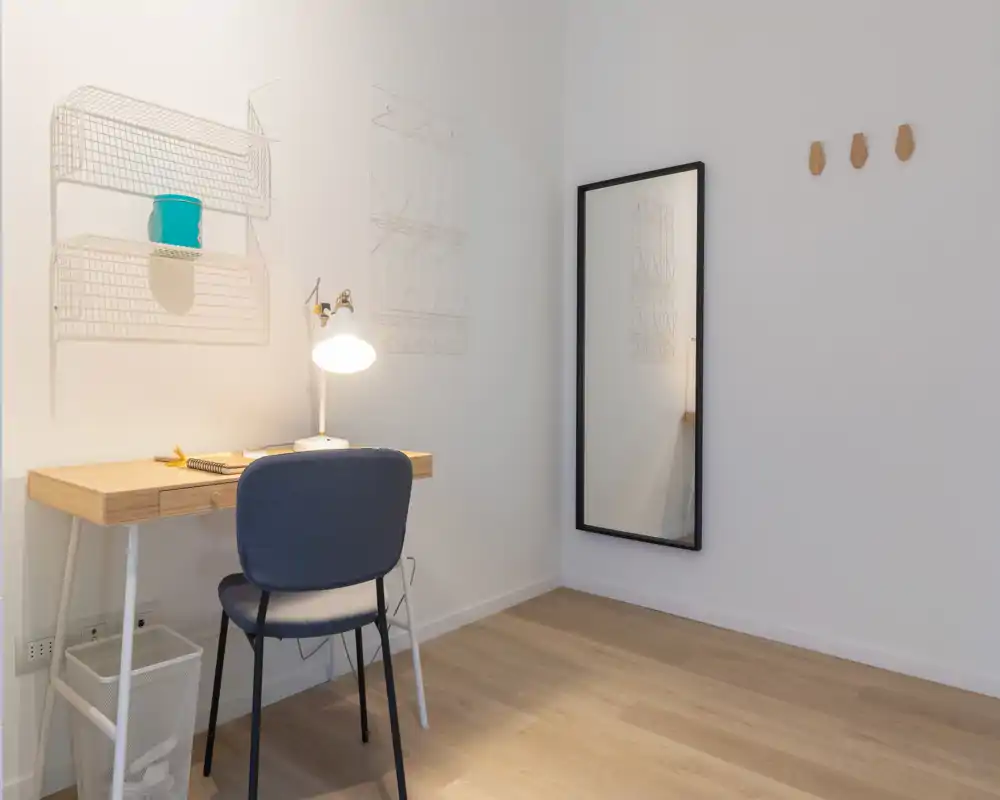 Rincón de sala minimalista y moderno con un pequeño escritorio de madera, silla y un espejo apoyado en una pared blanca; espacio luminoso y despejado, ideal para trabajar o relajarse.