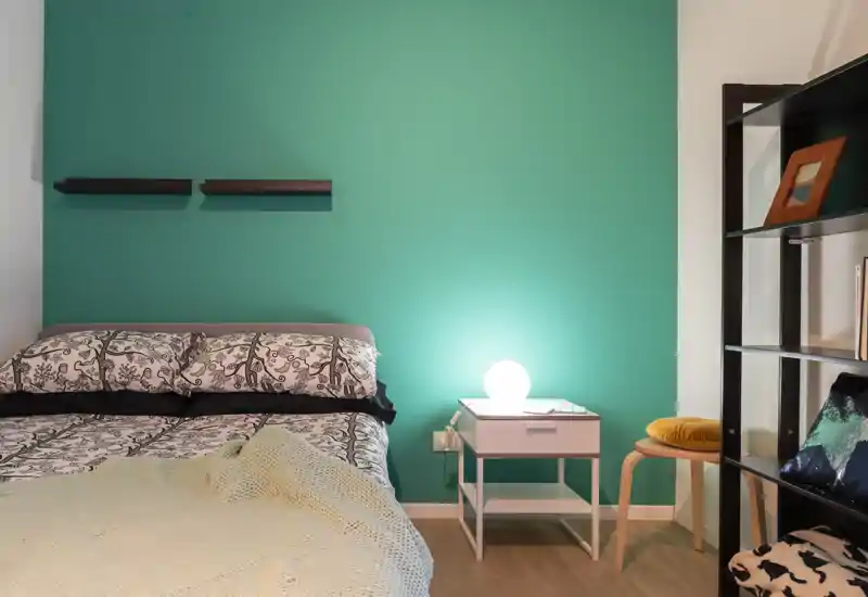 Dormitorio bien decorado con cama doble estampada frente a una pared turquesa, mesita con lámpara luminosa y estantería ordenada — acogedor y moderno.