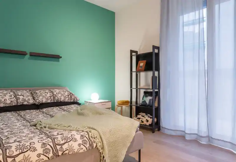 Bright, modern bedroom with a patterned bed, soft throw blanket and a bedside lamp; large sheer curtains let in natural light creating a calm, inviting atmosphere.