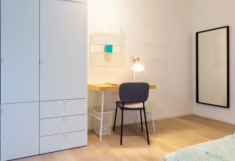 Bright, minimalist bedroom corner with a bedside/desk area, chair, wardrobe and partial bed in frame — clean wooden floor and a full-length mirror enhance the airy feel.