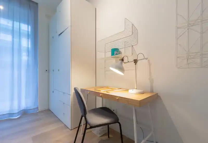 Small, modern bedroom corner with a wooden desk, task lamp and built-in wardrobe beside a sheer-windowed wall; clean lines and soft natural light create a calm workspace.