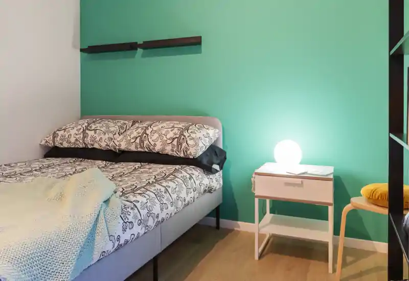 Dormitorio acogedor y bien iluminado con cama doble estampada, manta de punto y mesita de noche minimalista con lámpara frente a una pared verde azulado.