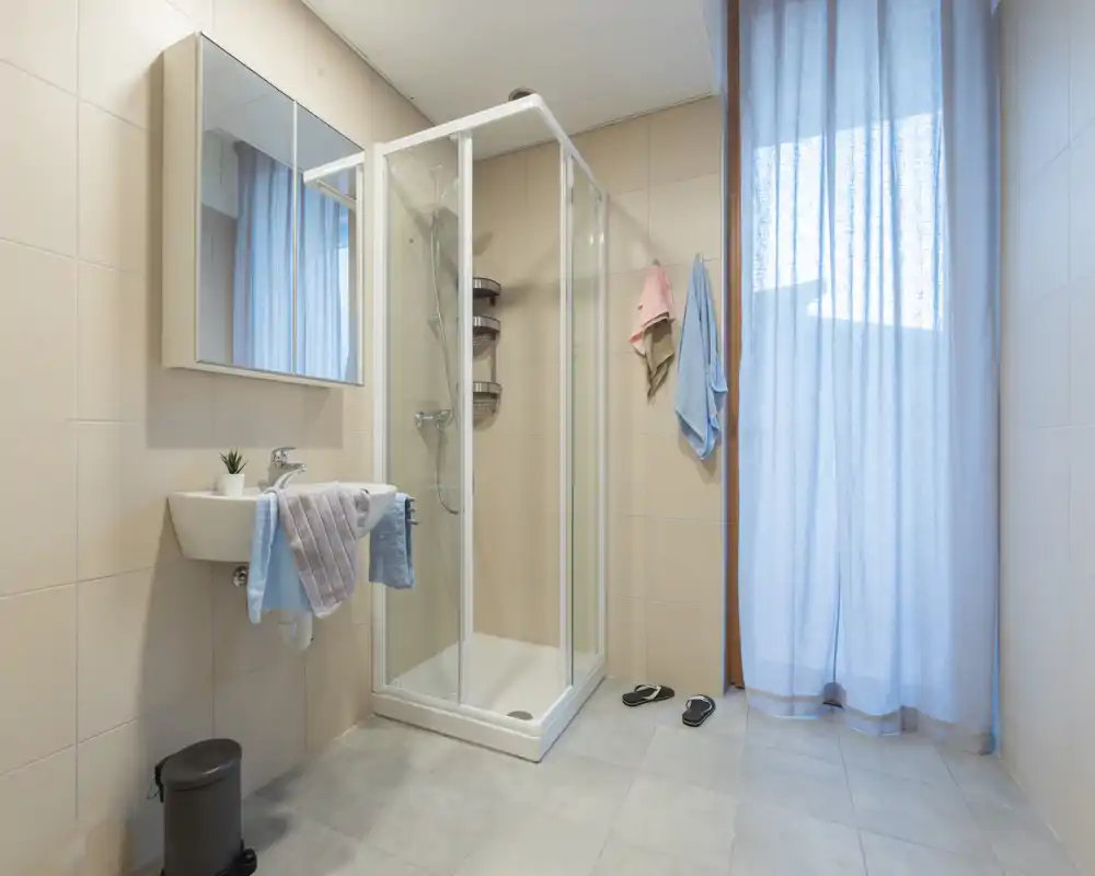 Baño moderno y luminoso con cabina de ducha de cristal, lavabo mural, espejo-armario y una gran ventana con cortina ligera.
