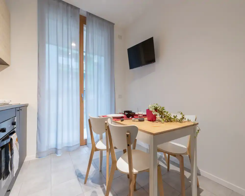 Pequeña zona de comedor con mesa de madera clara y cuatro sillas junto a una puerta de cristal; la mesa decorada y una planta crean un ambiente moderno y acogedor.