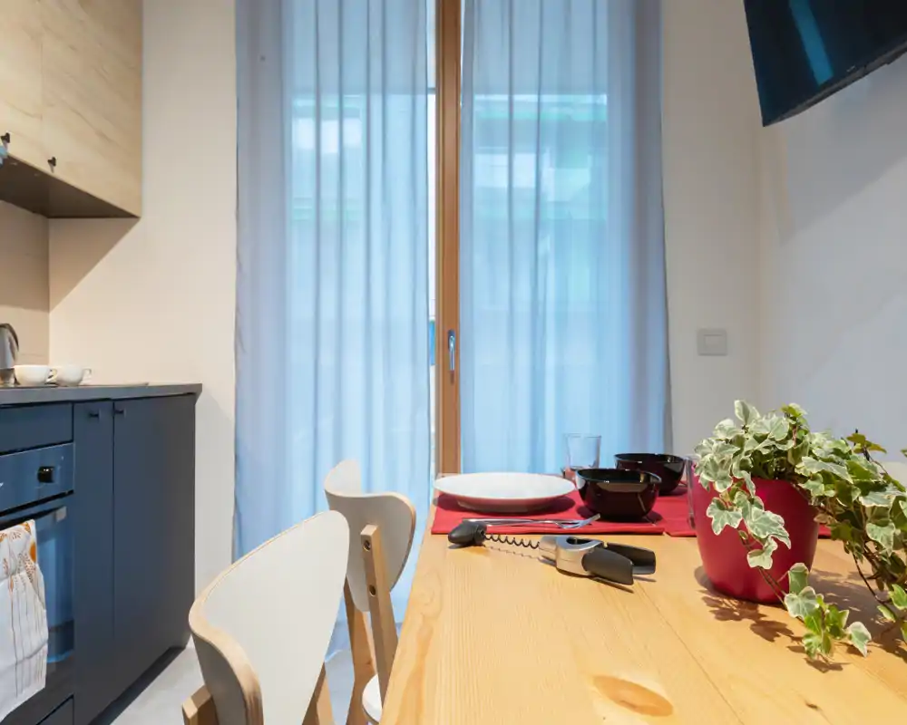 Cocina moderna con zona de comedor, mesa de madera puesta para dos, planta en maceta y luz natural desde la puerta de cristal.