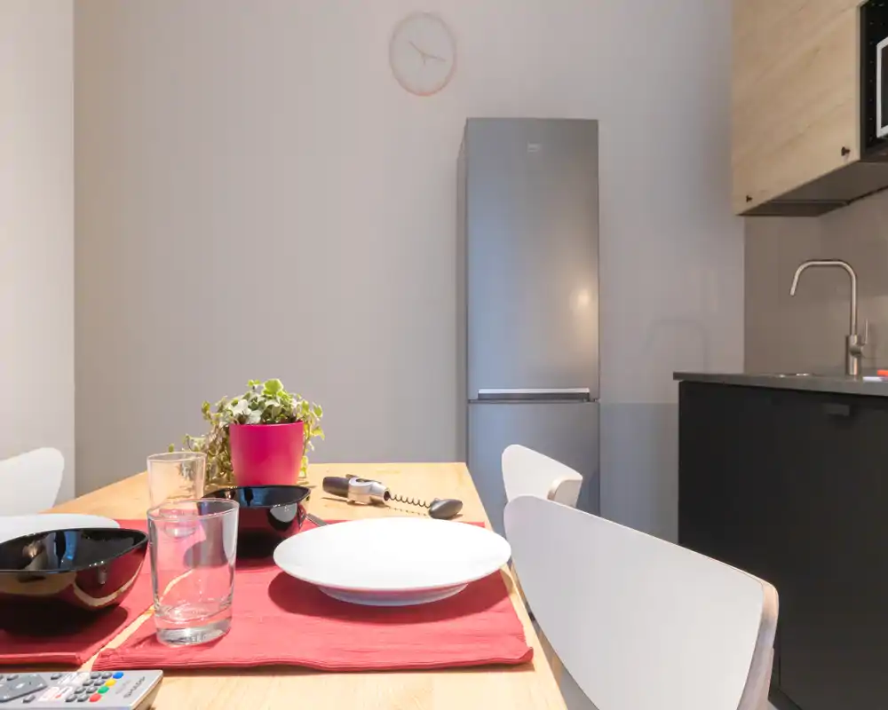 Cocina americana moderna y luminosa con mesa de madera puesta para comer, frigorífico visible y fregadero integrado — ideal para estancias urbanas pequeñas.