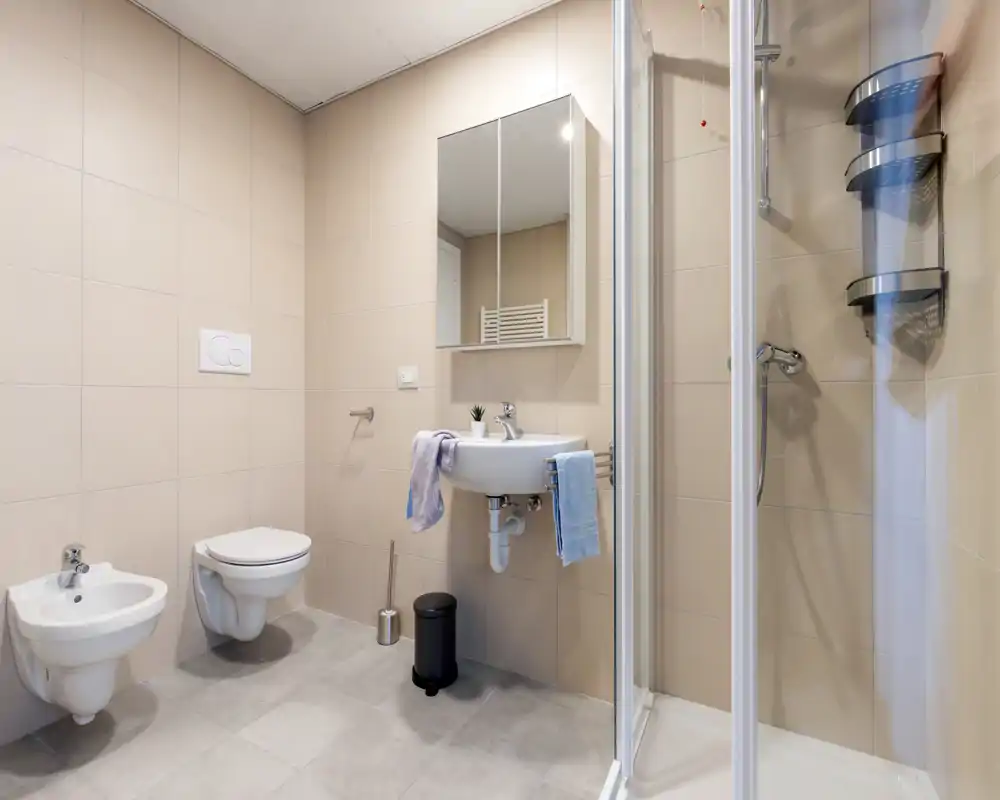 Baño moderno y bien iluminado con inodoro suspendido, bidé, lavabo con mueble espejo y cabina de ducha de cristal; azulejos beige y accesorios ordenados crean un ambiente limpio y acogedor.