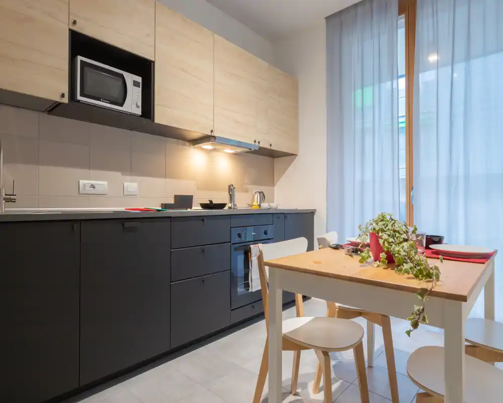 Cocina moderna equipada con armarios superiores de madera y muebles inferiores oscuros, horno y microondas integrados, pequeña mesa de madera con sillas y planta junto a una ventana luminosa.