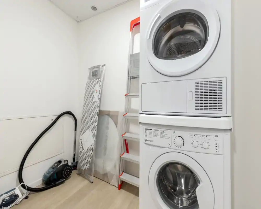 Cuarto de lavado/utility compacto con lavadora y secadora apiladas, aspiradora, tabla de planchar y escalera; espacio limpio, luminoso y funcional.