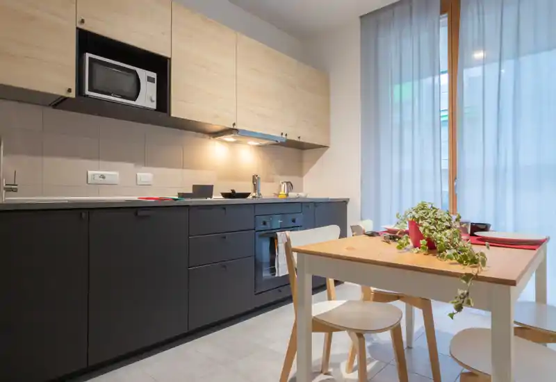 Modern, compact kitchen with matte dark lower cabinets, light wood upper cabinets, built-in oven and microwave, and a small dining table set by a bright window — styled with a plant and place settings for a welcoming feel.