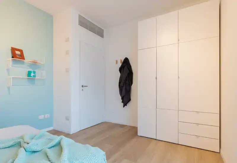 Bright, minimal bedroom corner with a visible portion of the bed, white wardrobe and pale blue accent wall. The image highlights storage and clean Scandinavian-style finishes.