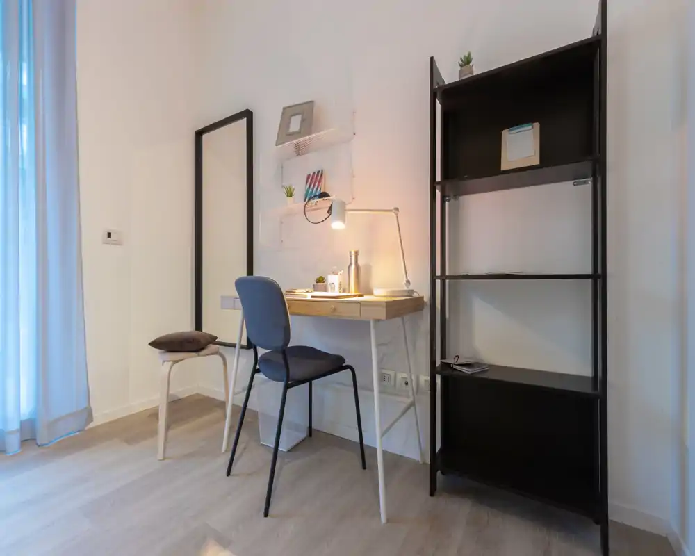 A tidy living area with a compact desk and chair set beside a tall shelving unit; soft daylight and a desk lamp create a warm, functional corner ideal for working or reading.