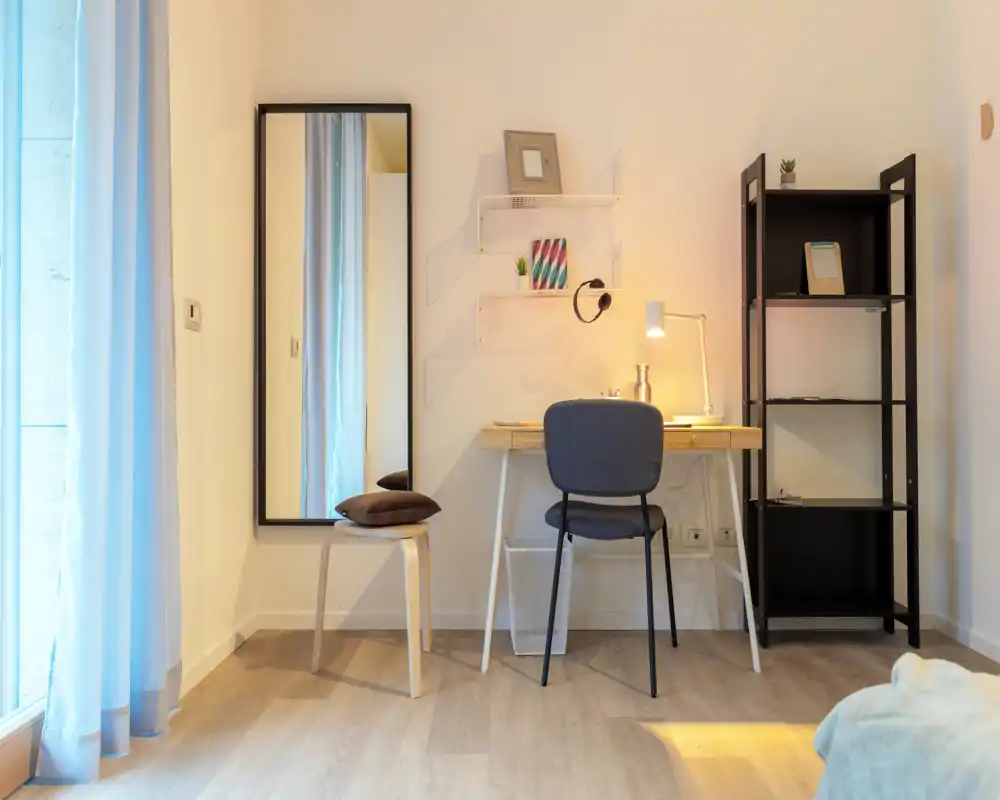 Modern, softly lit bedroom corner with a small desk, chair and shelving; a bed corner is visible in the foreground. Ideal for a compact, well-styled rental room with workspace and natural light.