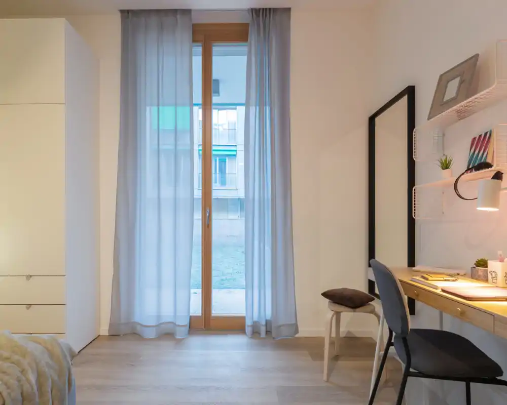 Bright, minimal bedroom corner with a sheer-curtained door, wardrobe and a tidy desk — clean lines and natural light create a calm workspace and sleeping area.