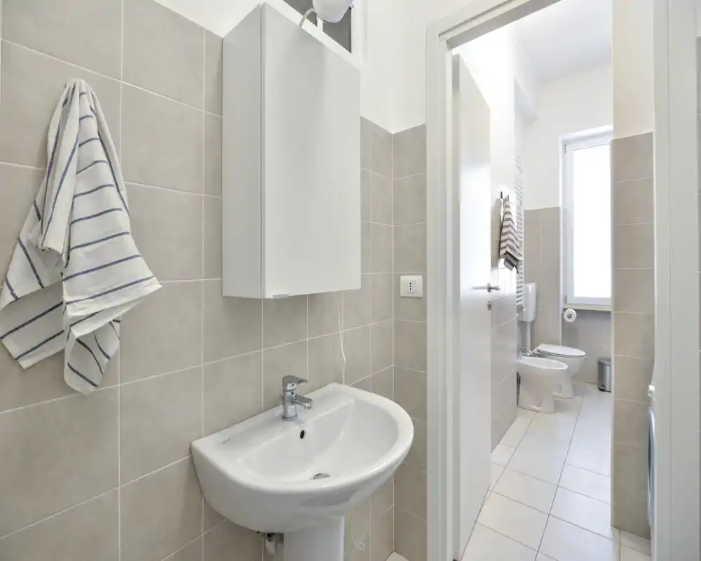 Baño moderno y luminoso con lavabo de pie, mueble de pared y zona de inodoro separada visible a través de la puerta.