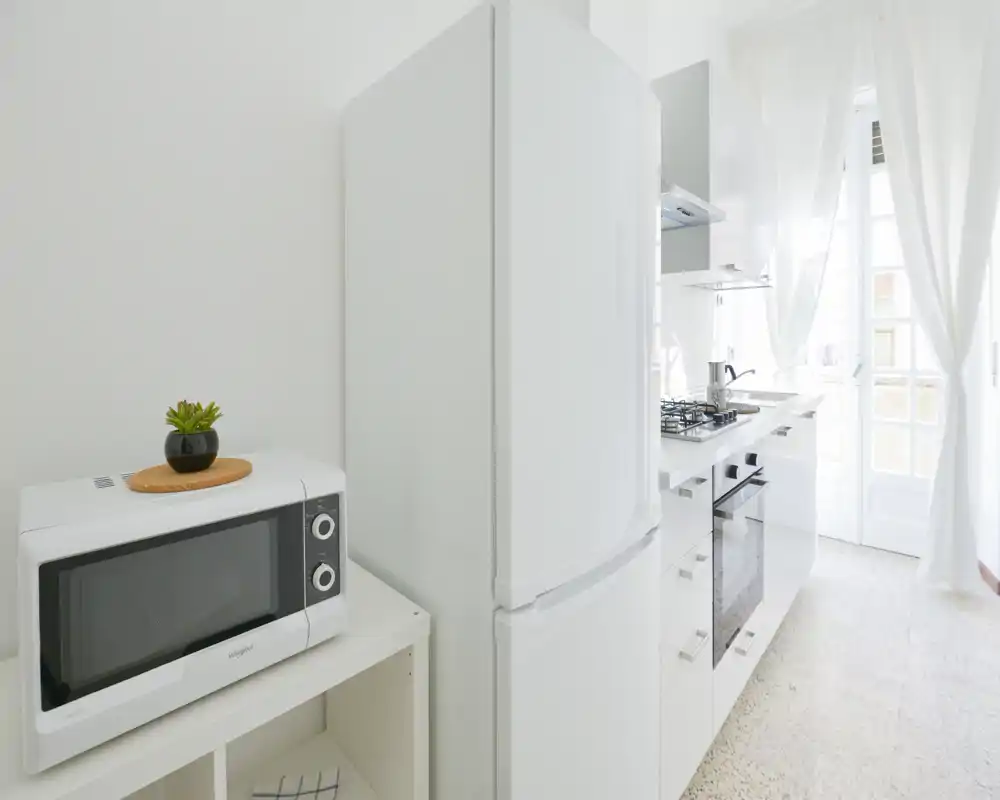 Cocina moderna y luminosa con muebles blancos, frigorífico independiente, horno empotrado y placa de gas; la luz natural entra a través de cortinas traslúcidas aportando sensación de amplitud.
