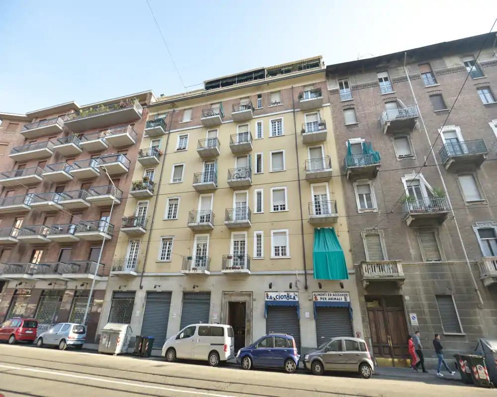 Fachada a pie de calle de un edificio residencial de varias plantas con pequeños balcones, locales en planta baja con persianas bajadas y coches aparcados en la vía.