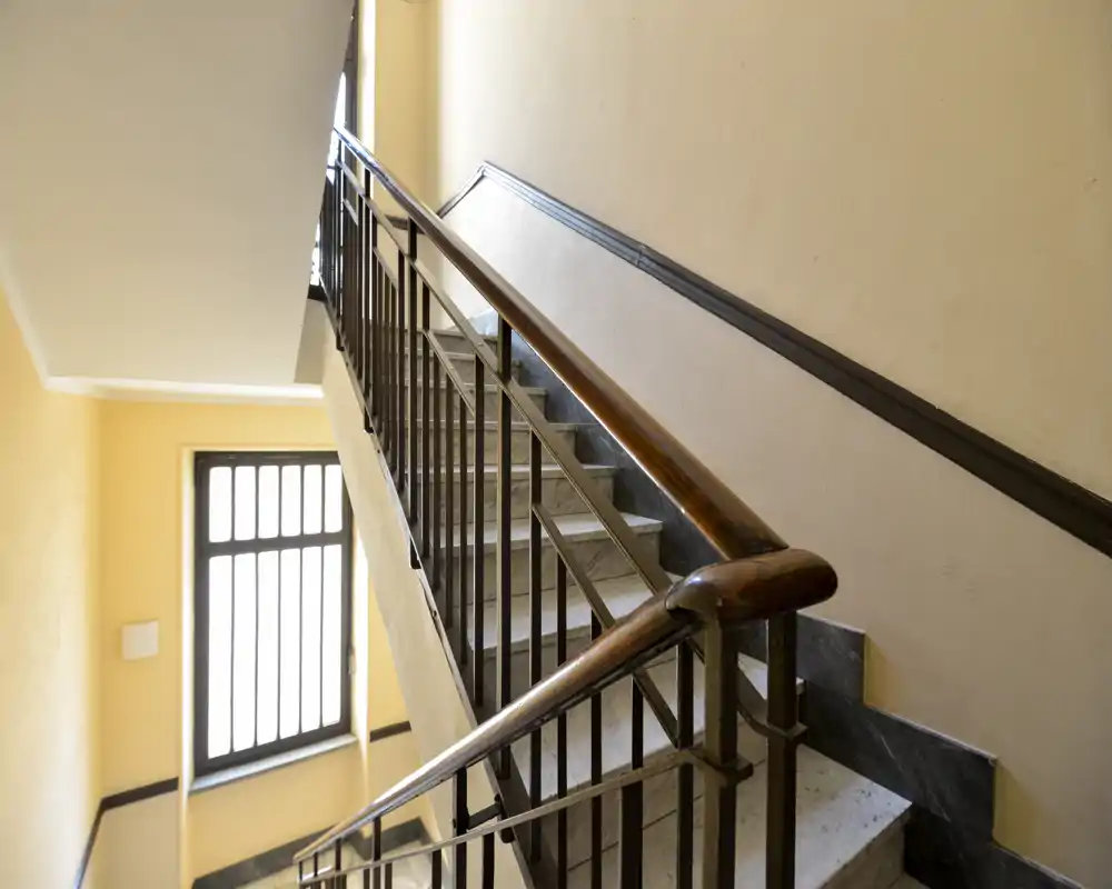 Tramo de escalera y entrada bien iluminados con pasamanos de madera y barandilla metálica; ventana brillante aporta luz natural y buena visibilidad de los peldaños.
