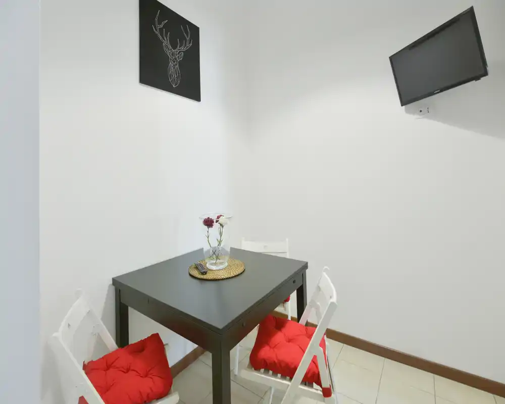 Rincón comedor compacto con mesa cuadrada oscura, tres sillas plegables blancas con cojines rojos y TV en la pared — ambiente luminoso y minimalista, ideal para apartamentos pequeños.