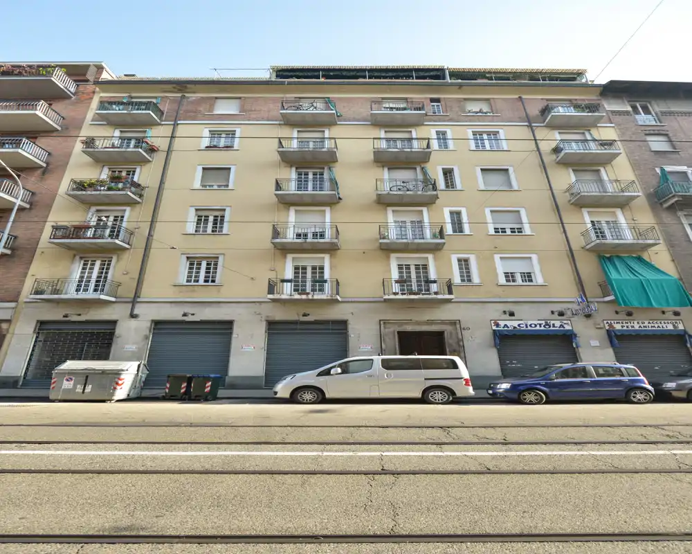Vista exterior de la fachada de un edificio residencial con balcones, persianas comerciales en planta baja, coches aparcados y vías de tranvía. Adecuada para describir la fachada o la propiedad a pie de calle.