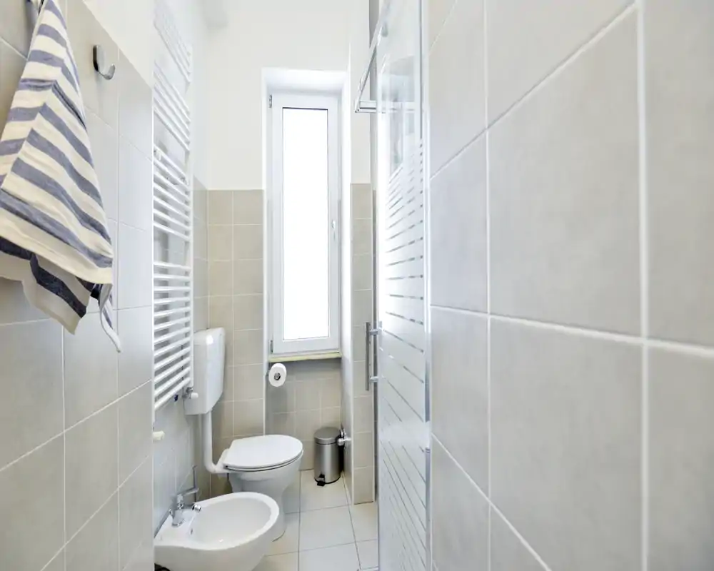 Baño compacto y luminoso con inodoro, bidé y cabina de ducha. Azulejos gris claro y una ventana estrecha que proporciona luz natural agradable.