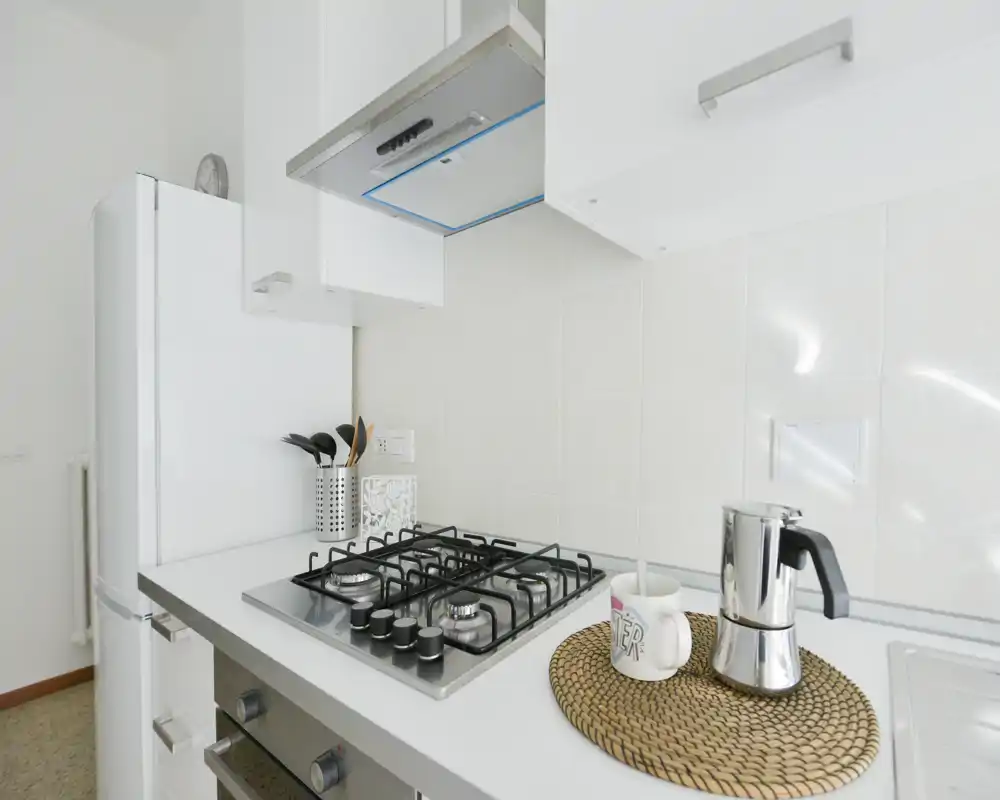 Rincón de cocina moderno y luminoso con placa de gas, campana y frigorífico; un pequeño salvamanteles de mimbre sostiene una cafetera moka y una taza, creando un aspecto limpio y minimalista.