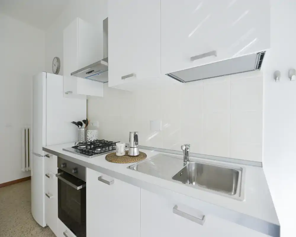 Cocina moderna y luminosa con muebles blancos, fregadero de acero, placa de gas y frigorífico; decorada con una moka y utensilios para una sensación limpia y acogedora.