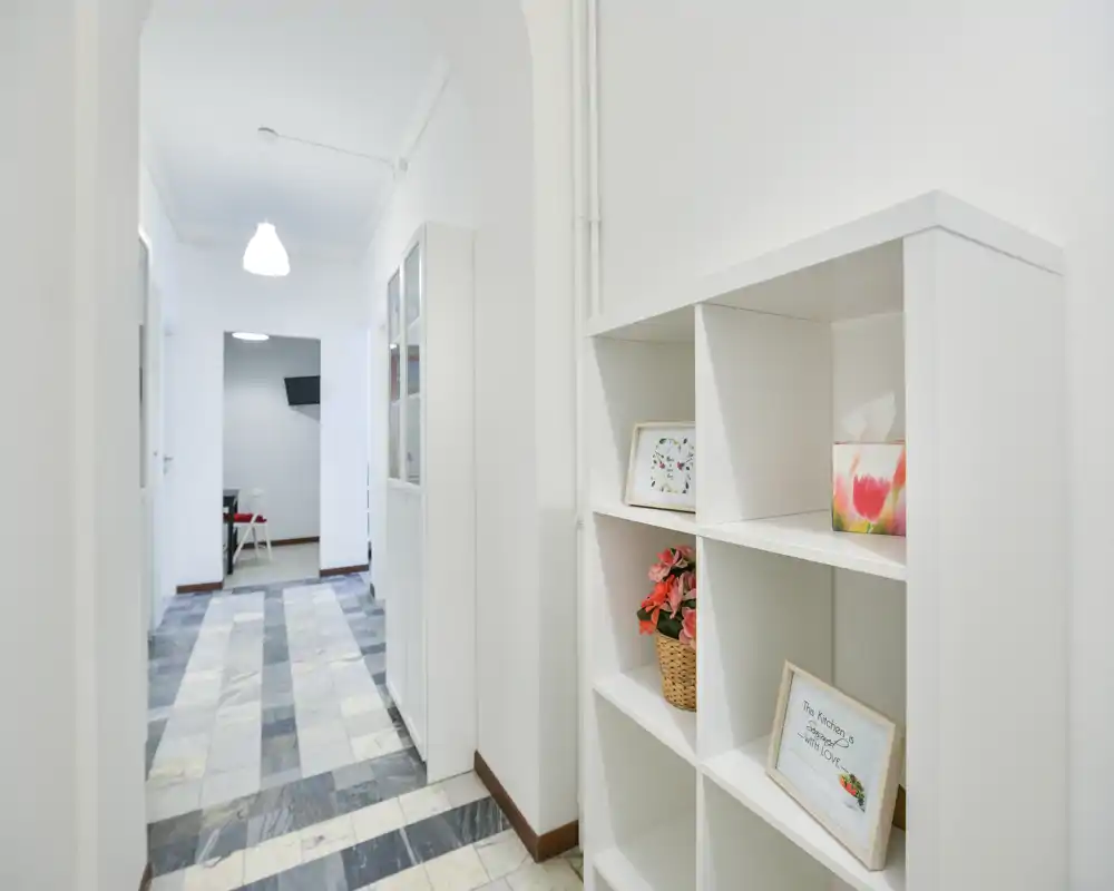 Entrada luminosa y limpia con paredes blancas, un arco y estanterías integradas; suelos de baldosas efecto mármol que conducen a una habitación interior.