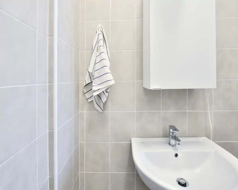 Rincón de baño moderno y compacto con lavabo de cerámica blanco, grifo cromado y una toalla a rayas colgada en la pared alicatada. Azulejos neutros y un pequeño armario con espejo ofrecen un aspecto minimalista y funcional.