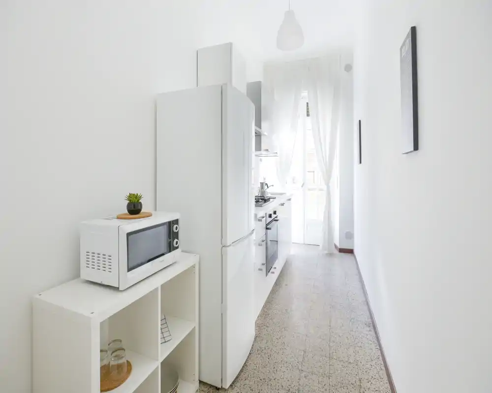 Cocina estrecha y luminosa con mobiliario blanco, nevera, horno y microondas; la luz natural a través de las cortinas traslúcidas crea un ambiente minimalista y aireado.
