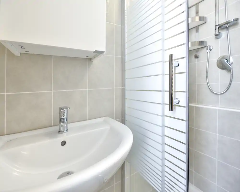 Baño compacto y moderno con lavabo de cerámica blanca y puerta de ducha de vidrio; azulejos grises y grifería cromada para un aspecto limpio y minimalista.