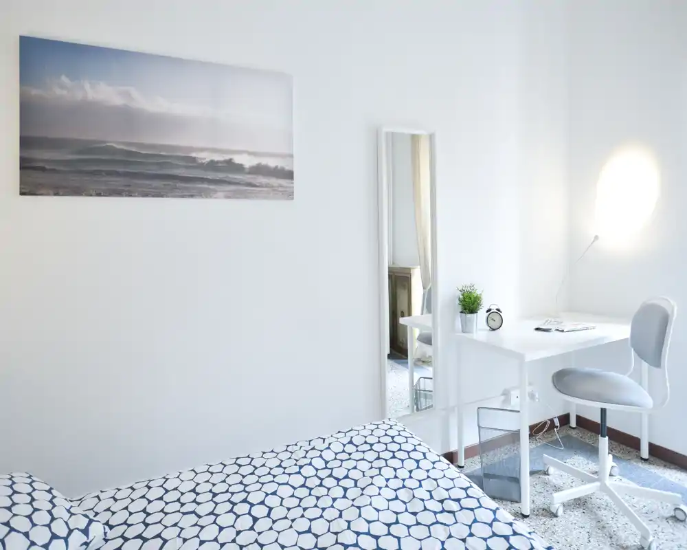 Chambre lumineuse et minimaliste avec couette à motif, petit bureau blanc avec chaise, miroir sur pied et tableau côtier — espace propre et aéré, idéal pour une personne.