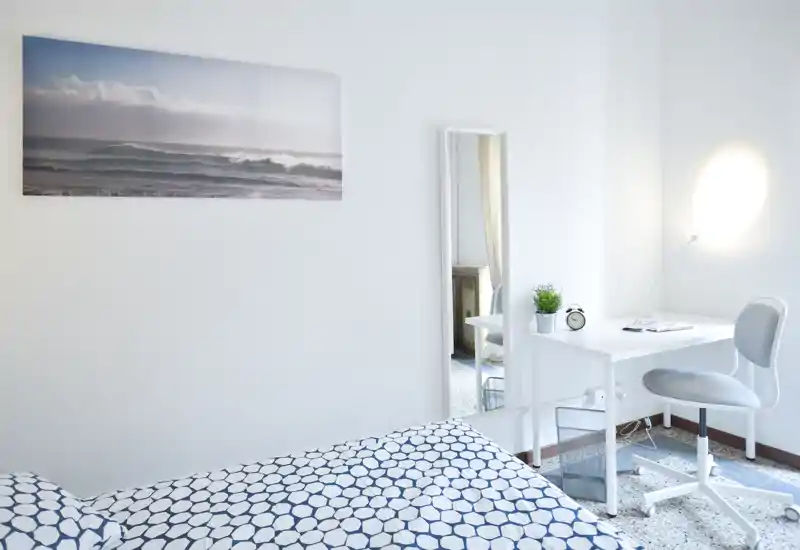 Chambre lumineuse et minimaliste avec couette à motif, petit bureau blanc avec chaise, miroir sur pied et tableau côtier — espace propre et aéré, idéal pour une personne.