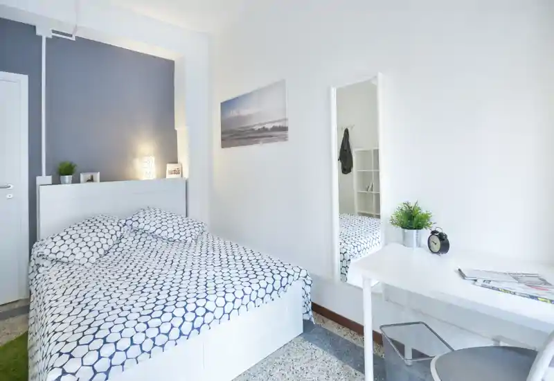 Chambre lumineuse et soignée avec un lit double en linge à motifs, une lampe de chevet et un bureau minimal avec miroir — idéale pour des annonces locatives mettant en avant confort et style moderne.