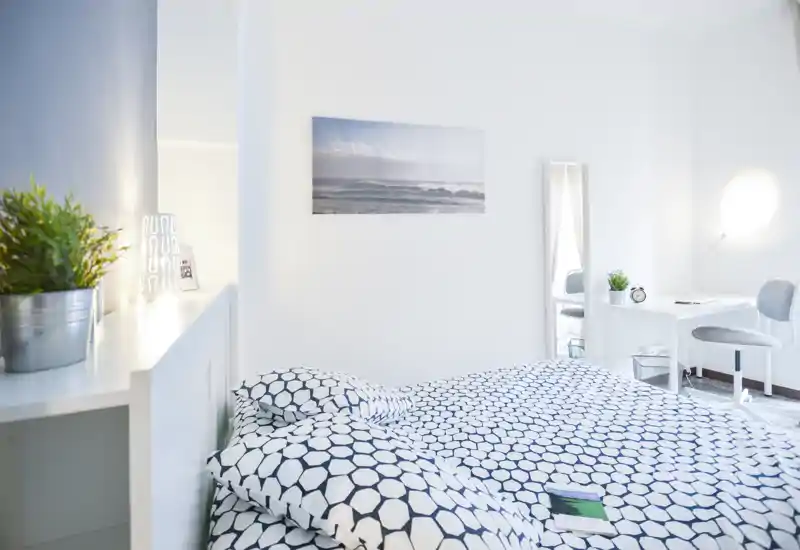 Chambre lumineuse et minimaliste avec une couette à motif, étagère de chevet avec plantes et lampe, et un petit coin bureau — bien éclairée et mise en scène pour des photos de location.