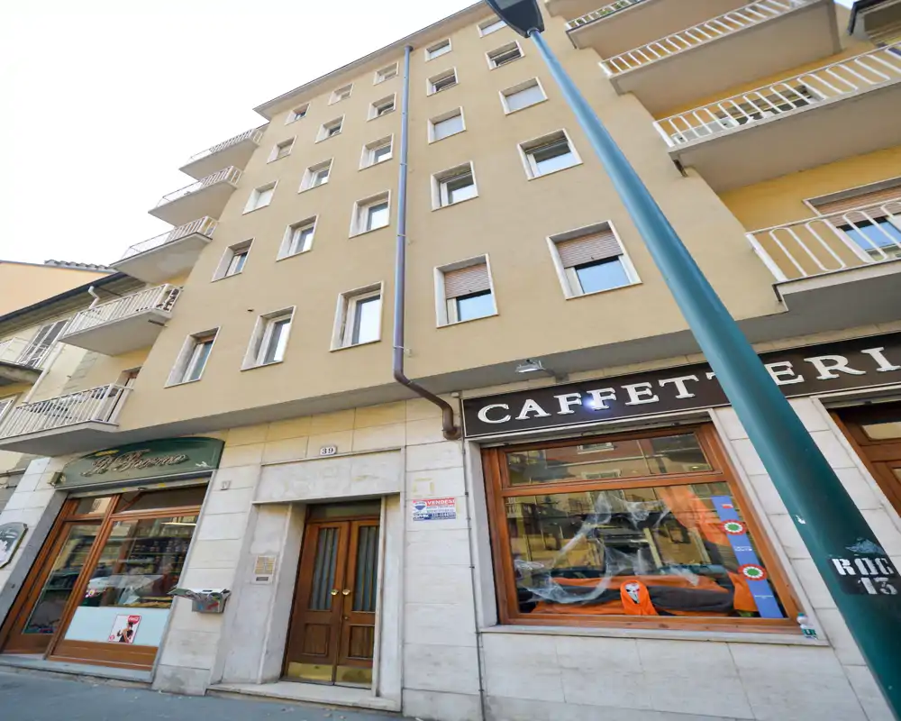 Vue extérieure d'un immeuble sur rue avec des commerces au rez-de-chaussée (dont une caffetteria). L'image montre la façade, les balcons et les portes d'entrée au niveau de la rue.