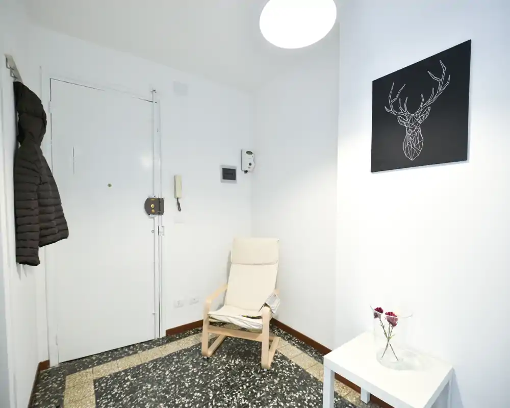 Petit hall d'entrée lumineux avec un fauteuil, un porte-manteau près de la porte et une table d'appoint avec des fleurs — espace d'accueil épuré et minimaliste.