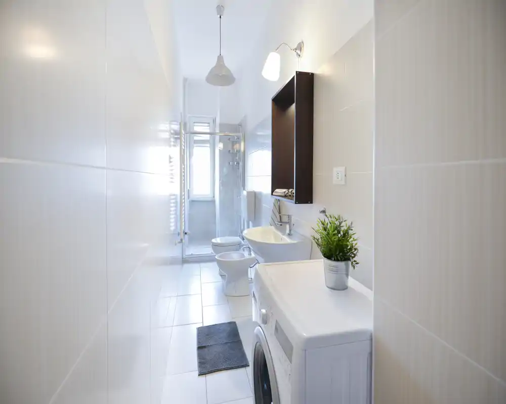 Salle de bains moderne et lumineuse avec carrelage blanc, douche vitrée, lavabo, bidet et lave-linge. Lignes épurées et lumière naturelle pour une ambiance fraîche et fonctionnelle.