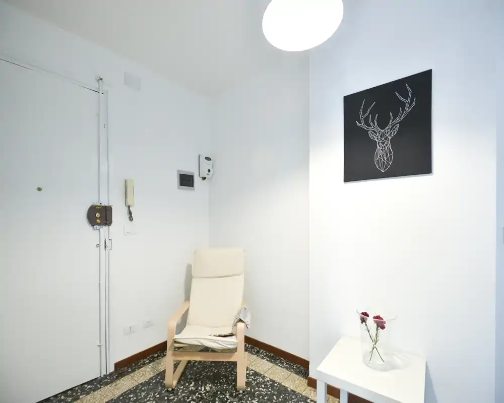 Entrée lumineuse et minimaliste avec un fauteuil, une petite table avec des fleurs et une affiche murale ; murs blancs et sol en terrazzo pour un accueil moderne et épuré.