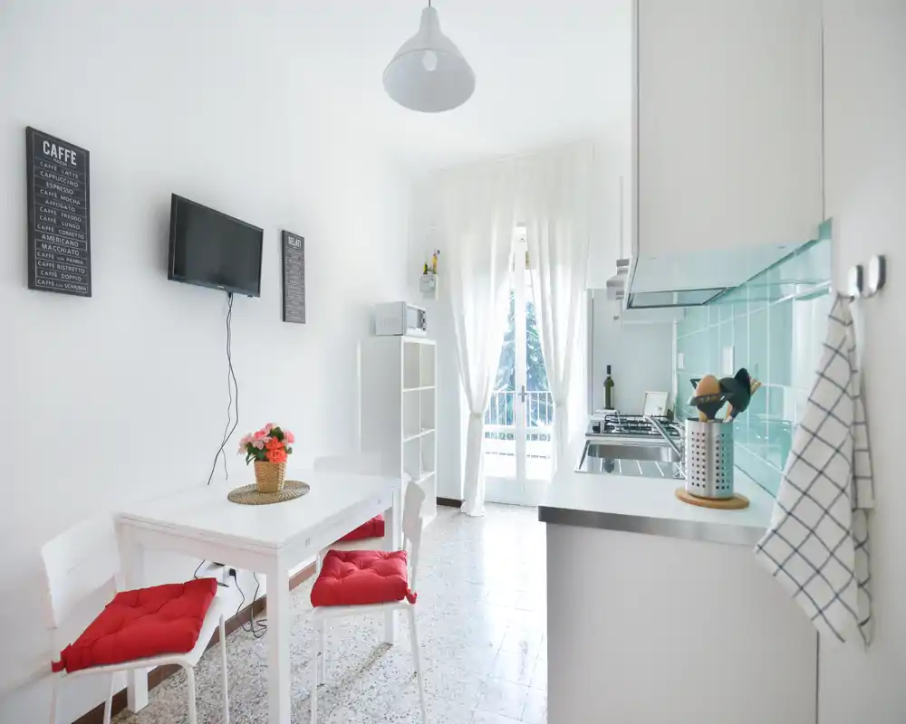 Kitchenette moderne et lumineuse avec une petite table à manger, des coussins rouges et une crédence carrelée. La lumière du balcon apporte une ambiance fraîche et aérée, idéale pour un studio ou petit appartement.
