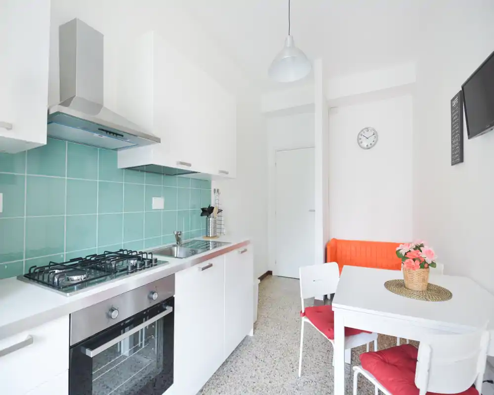 Cuisine compacte et moderne avec placards blancs, crédence en carrelage turquoise, four encastré et plaque à gaz. Une petite table avec chaises aux coussins rouges et un banc confortable rendent l'espace pratique et chaleureux.