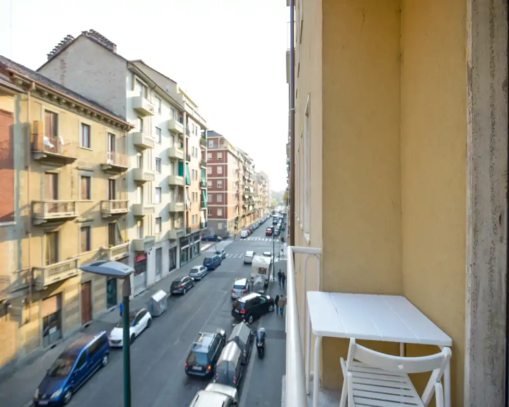 Petit balcon privé donnant sur une rue de la ville, avec table et chaise pliantes — vue urbaine lumineuse idéale pour le café du matin.