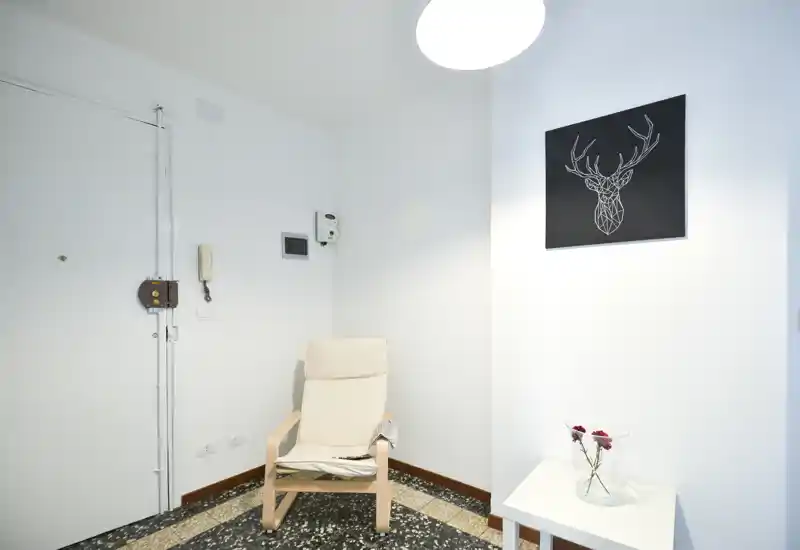 Ingresso luminoso e minimal con una poltrona, un piccolo tavolino con fiori e una stampa appesa; pareti bianche e pavimento in graniglia donano un aspetto moderno e ordinato.