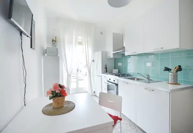 Cucina compatta e luminosa con pensili bianchi e rivestimento piastrellato turchese, dotata di forno, piano cottura a gas e lavello; piccolo tavolo da pranzo con fiori e luce naturale dalle porte-finestre.