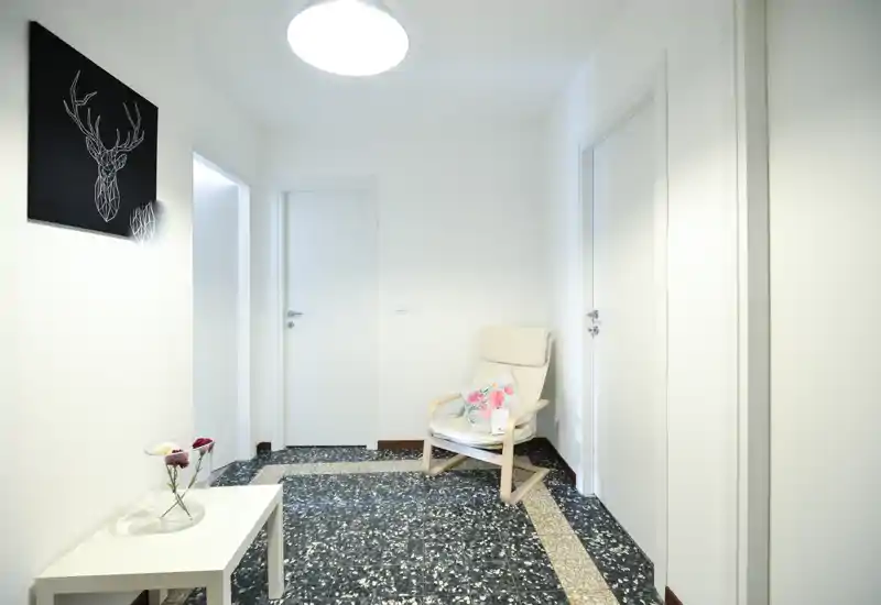 Ingresso luminoso e minimale con una poltrona chiara, piccolo tavolino con vaso e tre porte bianche — arredo pulito e moderno con pavimento in stile graniglia.
