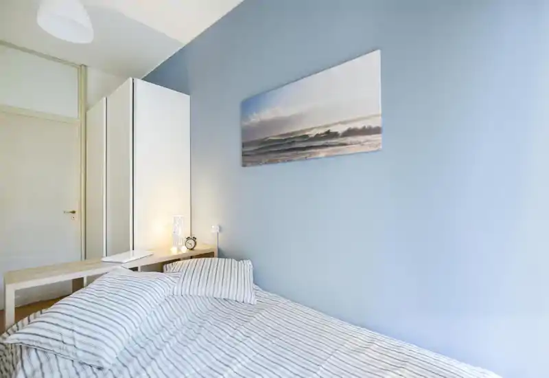 Bright, minimal bedroom with a neatly made bed, striped bedding and soft blue walls. A wardrobe and bedside table with a lamp and alarm clock complete the calm, coastal-inspired decor.